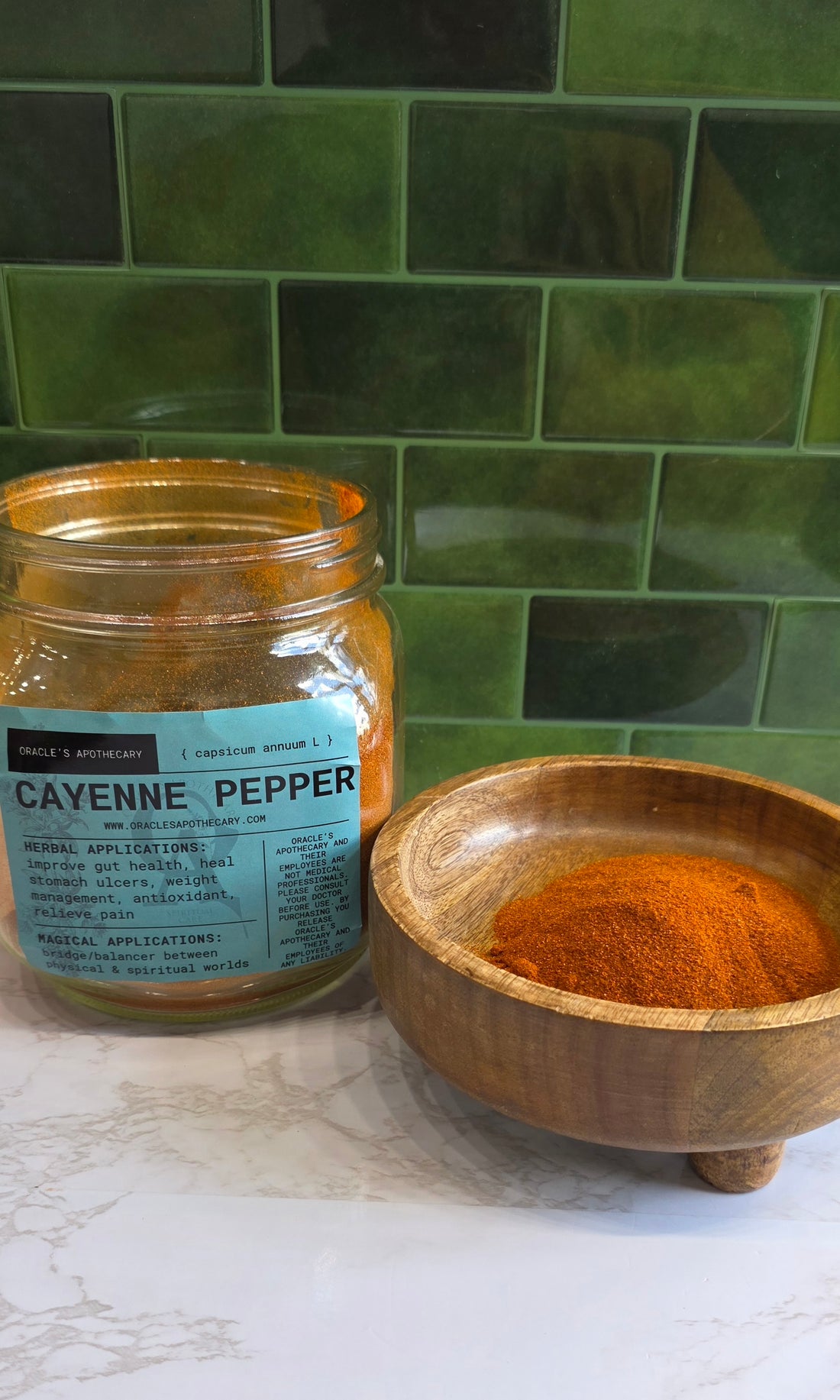 Cayenne Pepper Powder in clear jar with label and in wood bowl on a marble counter top with green tile background