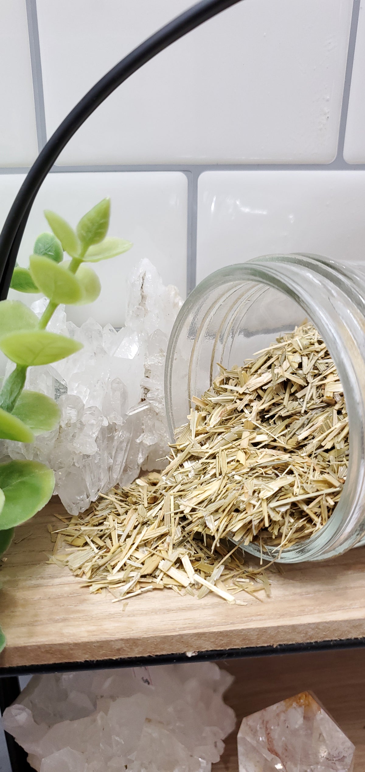 A glass jar on a wooden surface with what appears to be dried oat straw protruding from it. There are white tiles in the background and a green plant to the left.