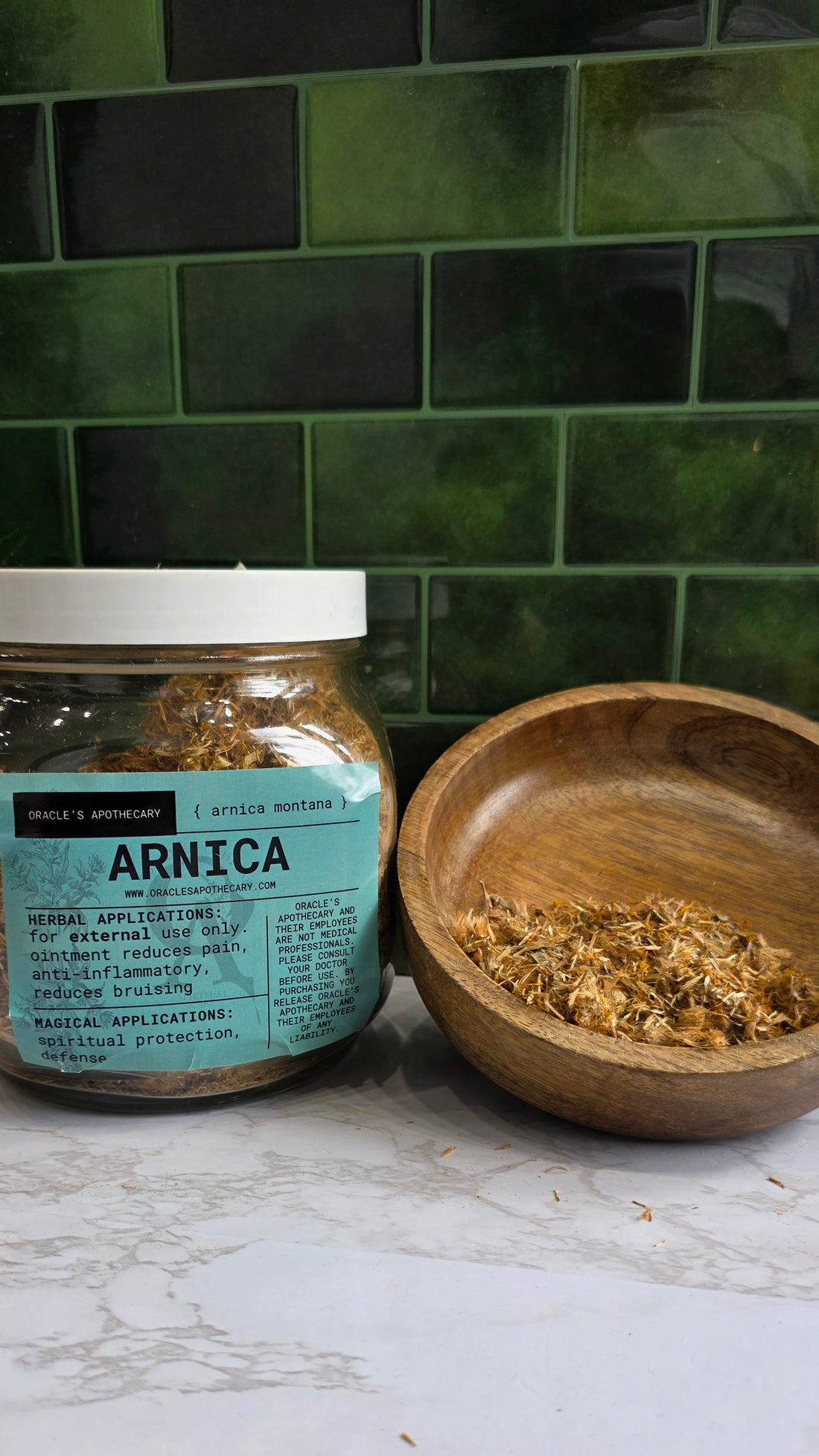 Arnica Flower in clear jar with label and in wood bowl on a marble counter top with green tile background 