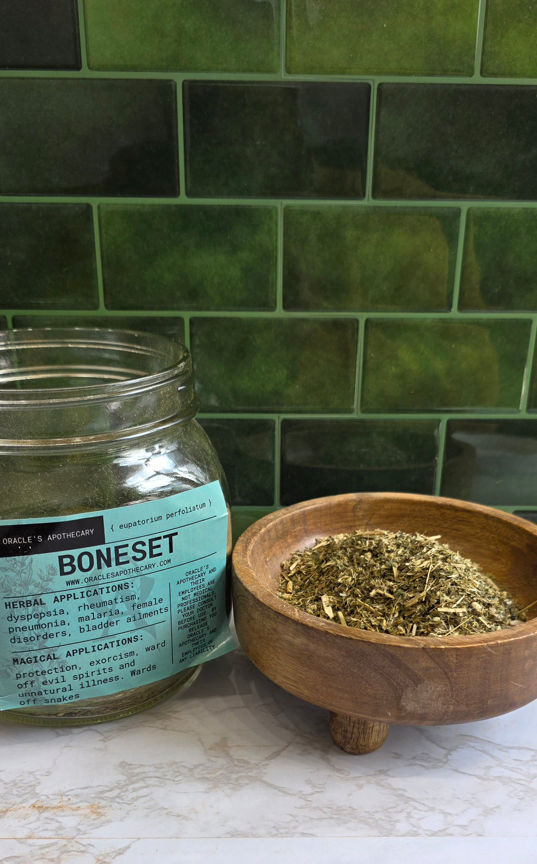 A clear glass jar labeled BONESET with a light blue paper label sits on a marble surface next to a small wooden bowl filled with dried green herbal leaves, set against a green subway tile background.