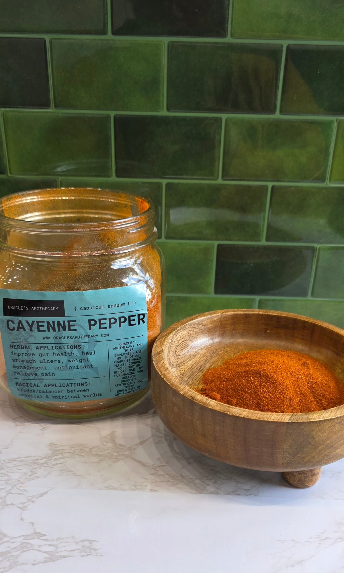 Cayenne Pepper Powder in clear jar with label and in wood bowl on a marble counter top with green tile background