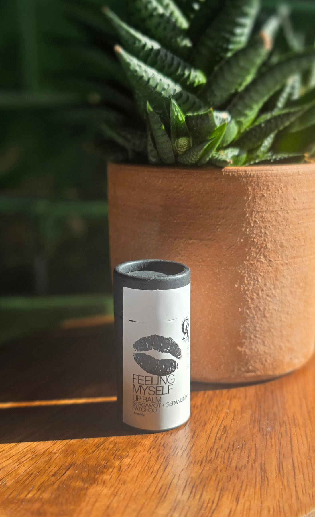 White label on black tube with black text and a black lid on a wooden surface with a plant in the background