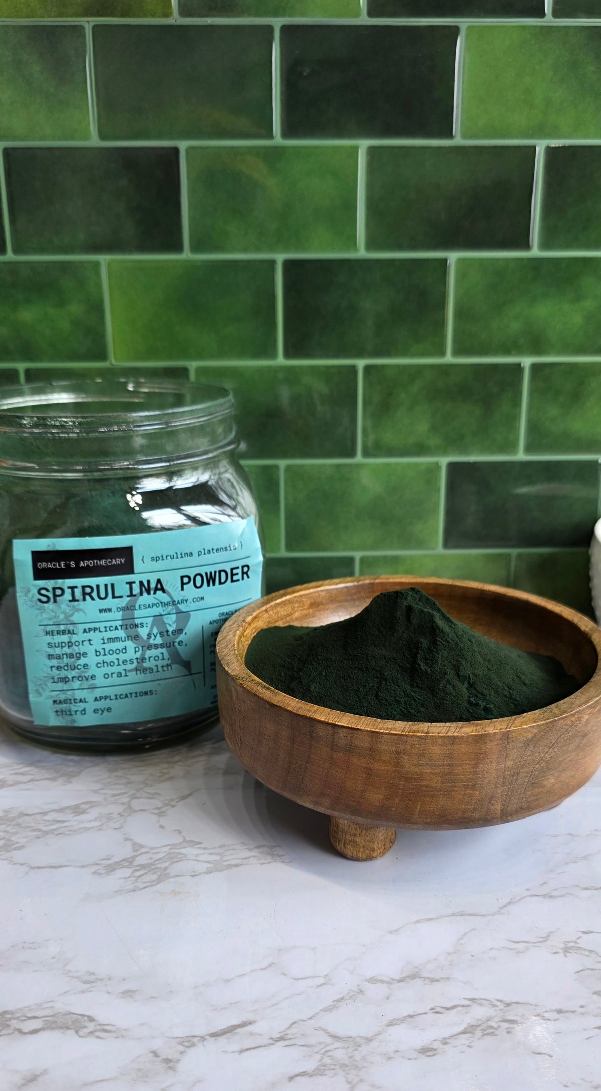 Organic Spirulina Powder in clear jar with label and in wood bowl on a marble counter top with green tile background 