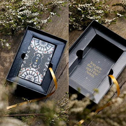 Card in a black box with a gold ribbon on a wooden surface with flowers.