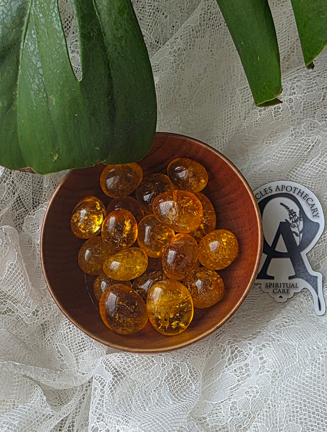 Brown bowl with amber-colored beads on a lace tablecloth with a plant leaf and logo in the background.